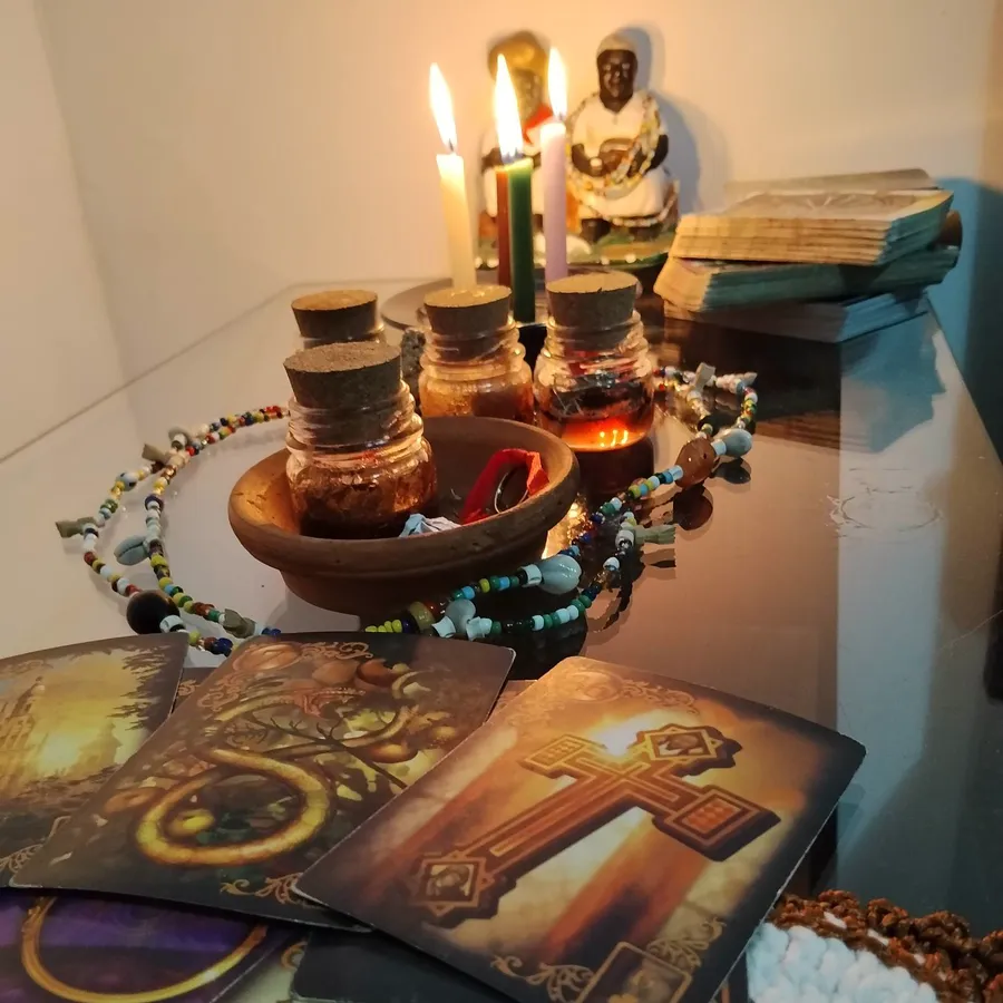 Mesa de atendimento da Taróloga Bianca com cartas abertas, vidros de preparo com rolha, guias de contas coloridas e velas diante de uma Preta Velha