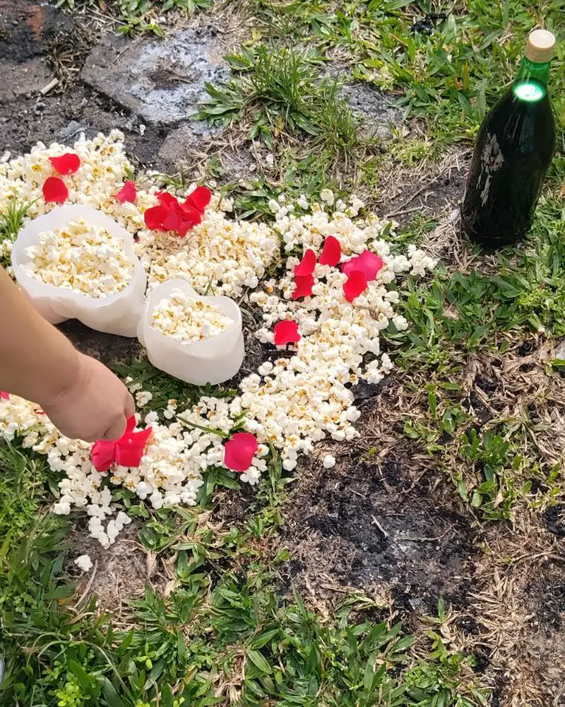 Mão colocando pétalas de rosa vermelha sobre uma oferenda de pipoca na grama, ao lado de uma garrafa