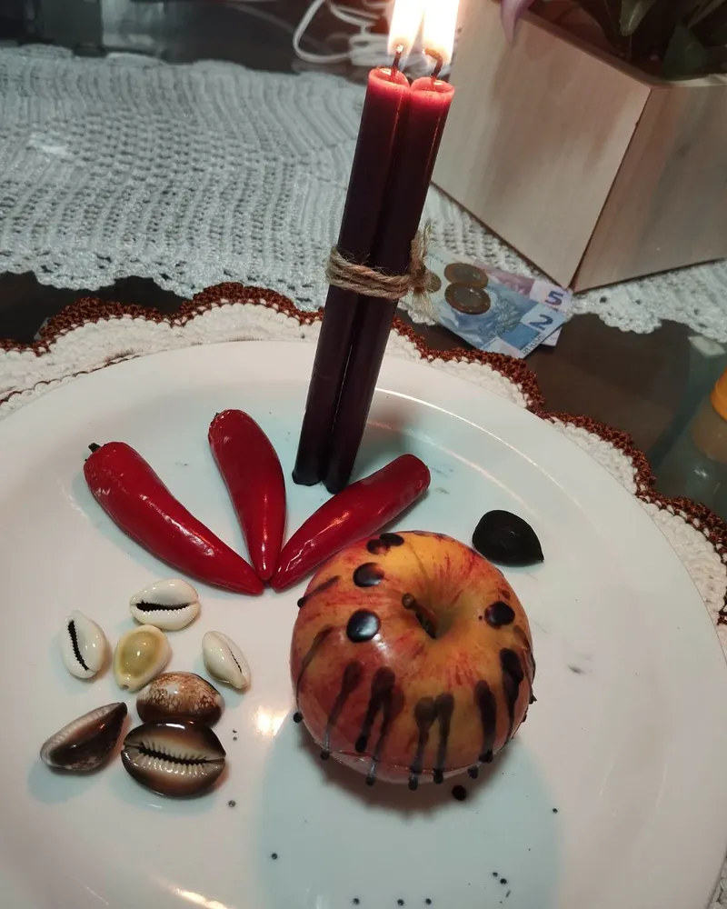 Prato branco com duas velas vermelhas amarradas e acesas, três pimentas vermelhas, búzios e uma maçã com cera escorrida