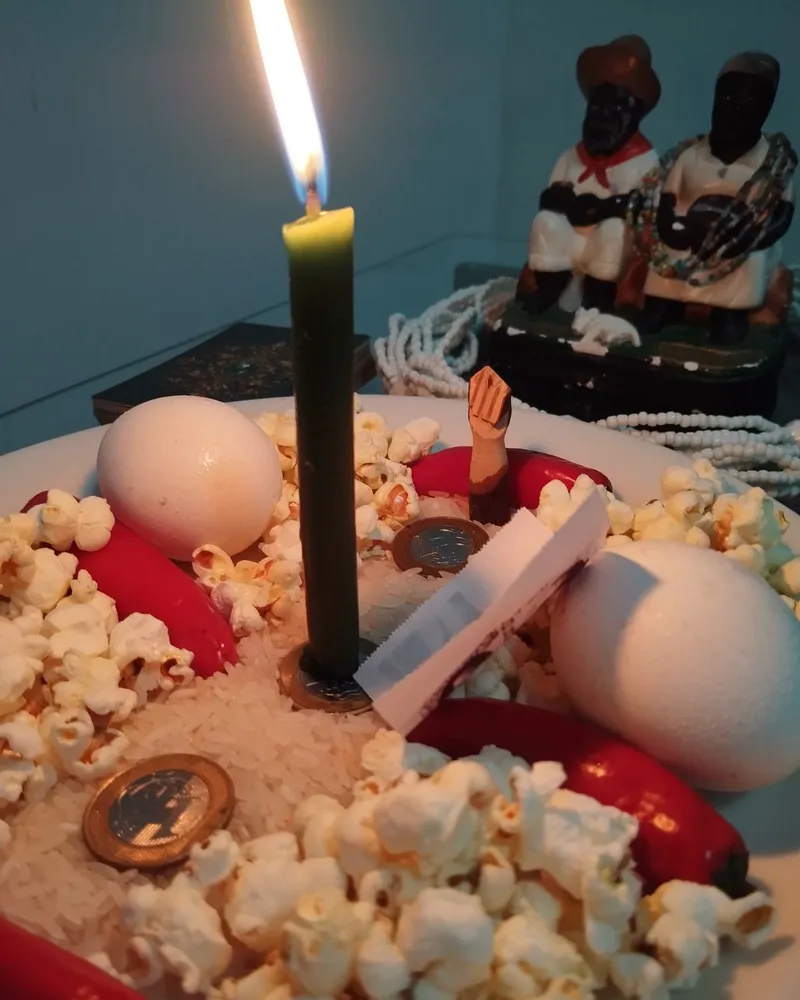 Vela verde acesa sobre arroz, com moedas, ovos, pimentas vermelhas e pipoca num prato branco, e os Pretos Velhos ao fundo