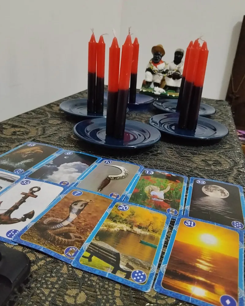 Velas vermelhas e pretas em pires azuis ao lado das imagens de Pretos Velhos, com cartas do baralho cigano abertas na mesa