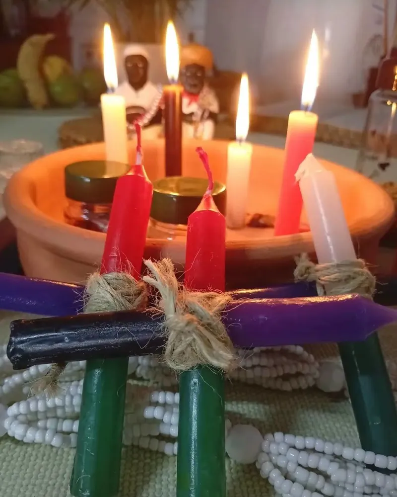 Velas brancas, vermelhas e rosa acesas dentro de um alguidar de barro, com velas verdes, roxas e pretas amarradas com barbante e guias de contas brancas na frente